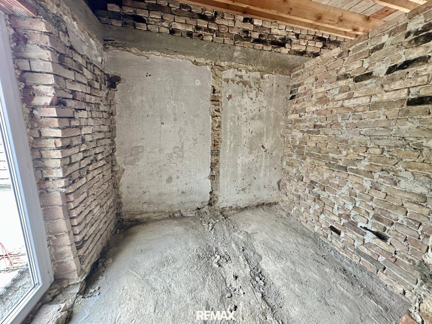 Rohbauzimmer mit freiliegenden Ziegelwänden und Betonboden, bereit für den Ausbau.