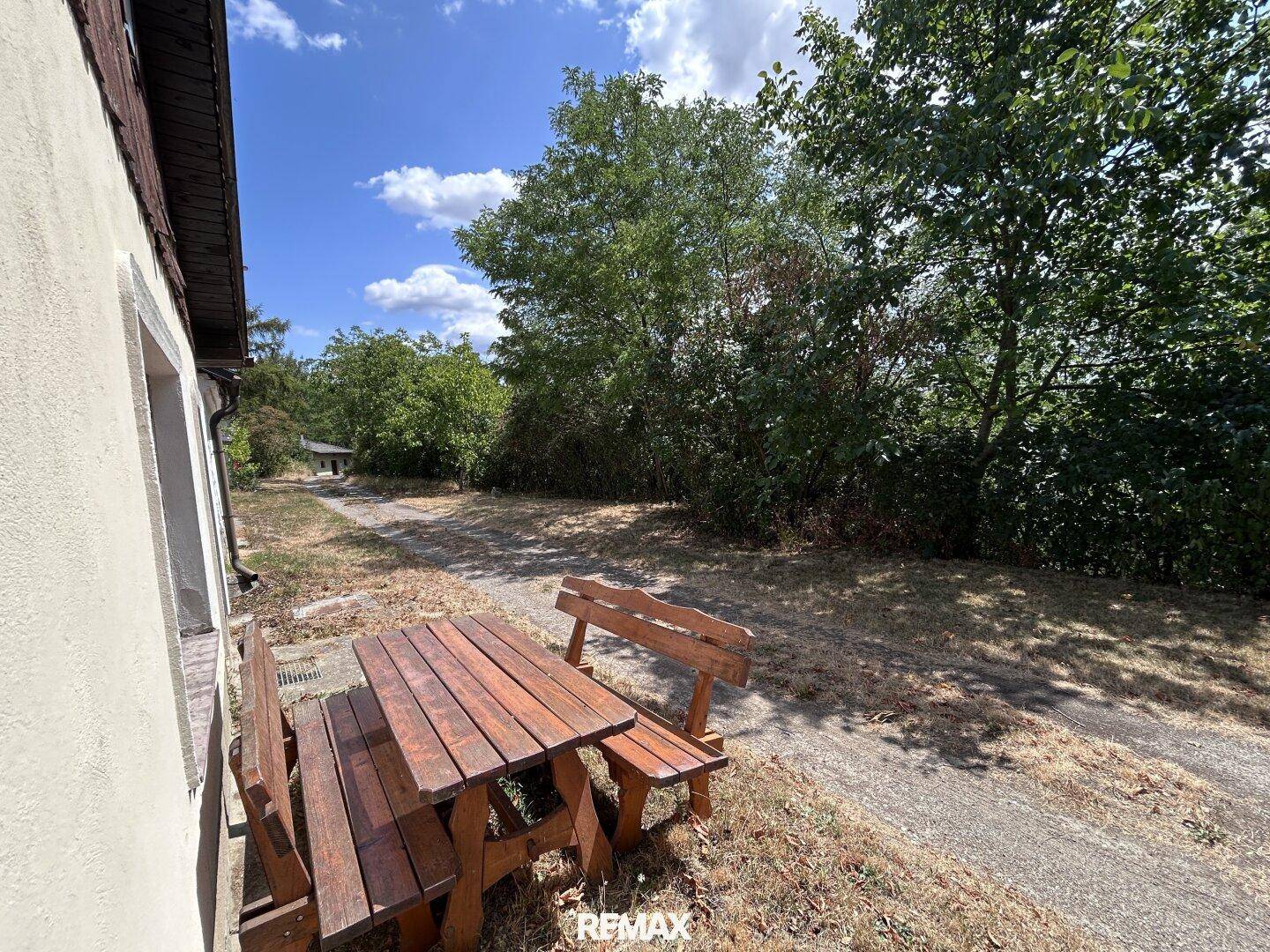 Seitlicher Blick auf das Haus mit Holztisch und Bänken im sonnigen Gartenbereich.