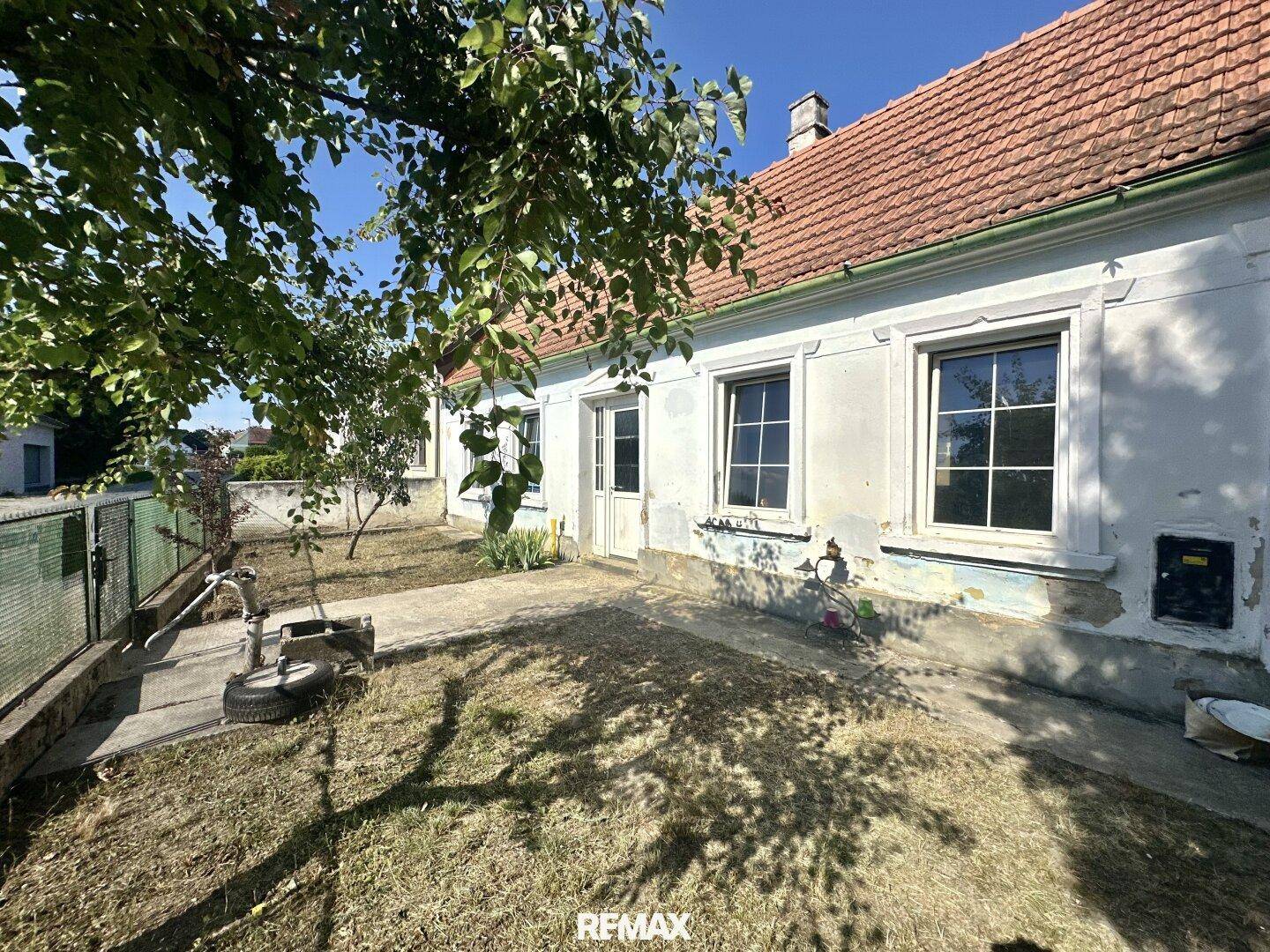 Vorderansicht eines alten Hauses mit Gartenbereich, Bäumen und einer manuellen Wasserpumpe.