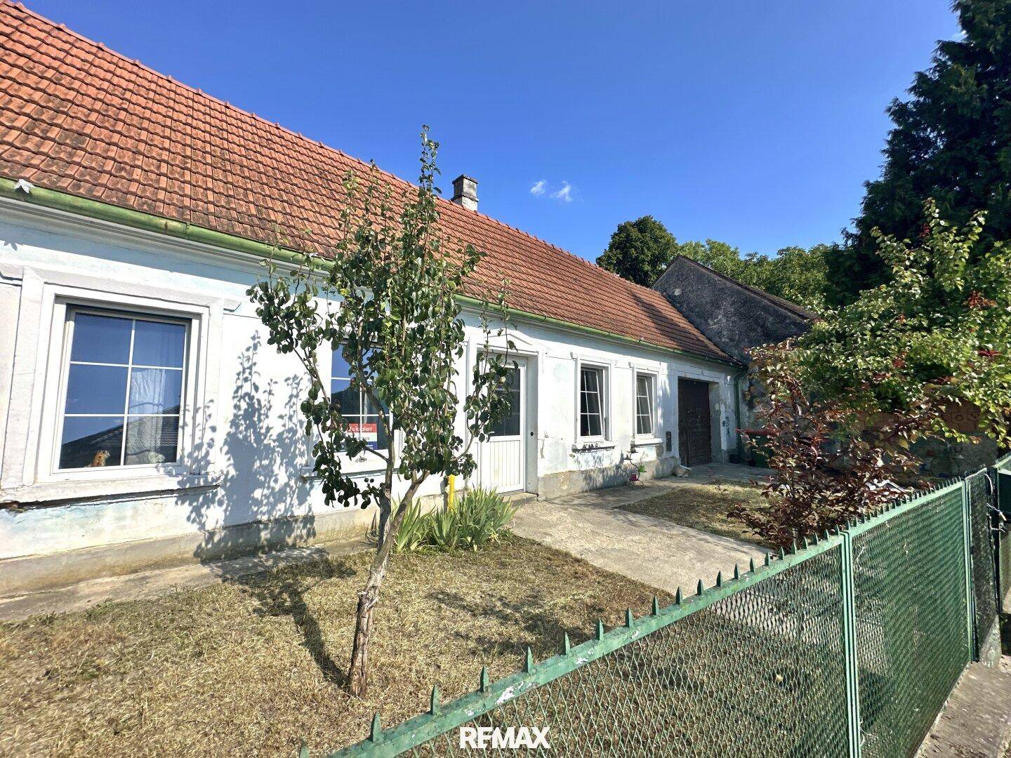 Ländliches Haus mit weißer Fassade, rotem Dach und einem kleinen Vorgarten unter blauem Himmel.