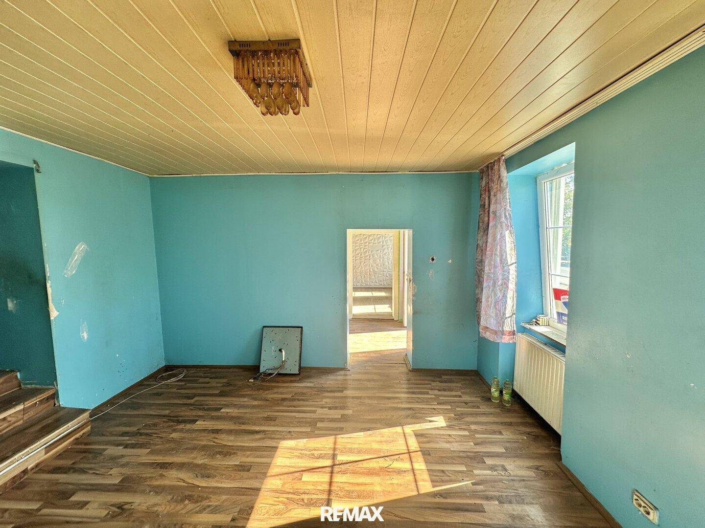 Wohnzimmer mit blauen Wänden, Holzdecke und Holzboden, mit Blick in den nächsten Raum.