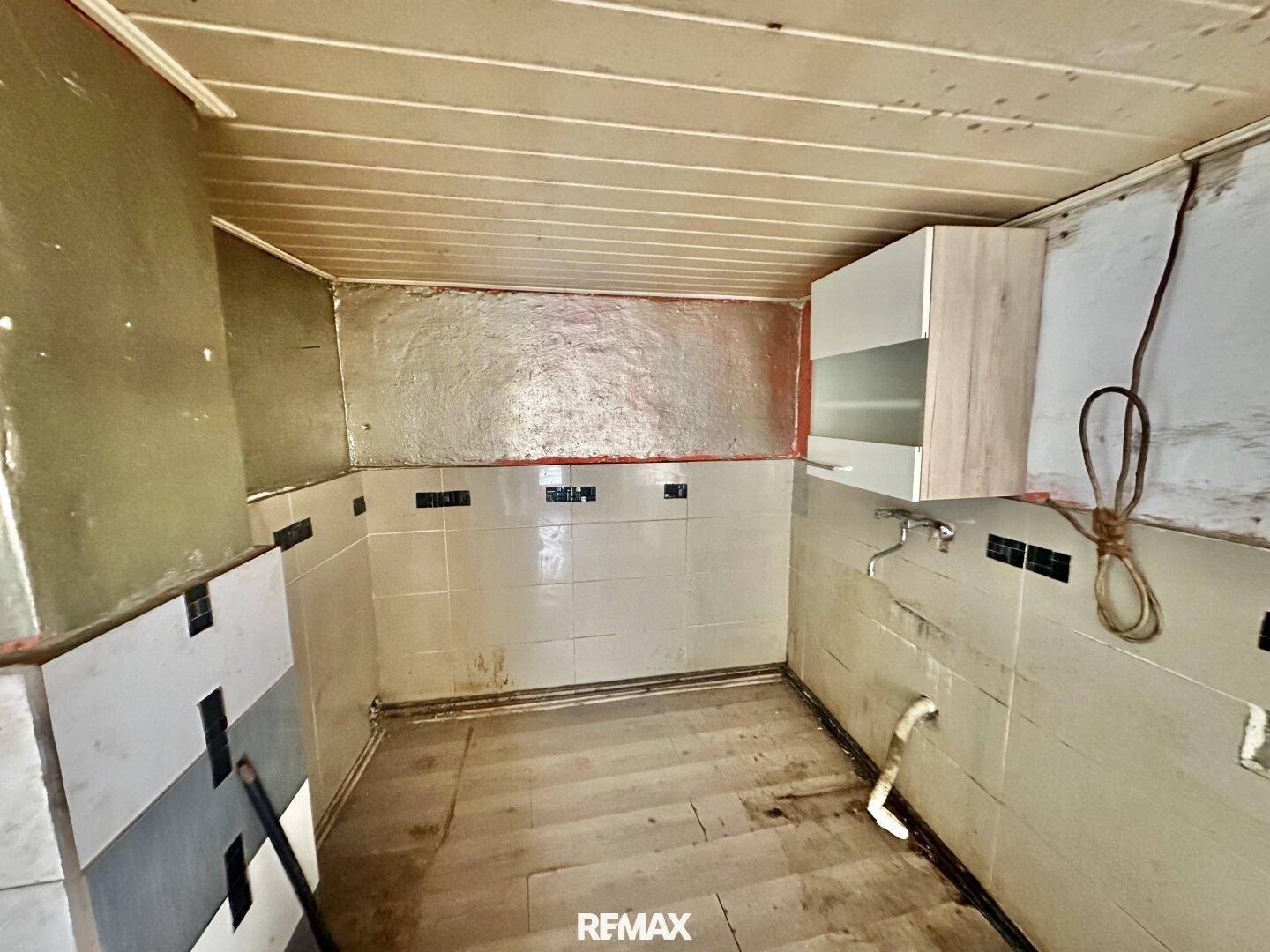 Küche in sanierungsbedürftigem Zustand mit alten Fliesen, Holzdecke und einem Hängeschrank.