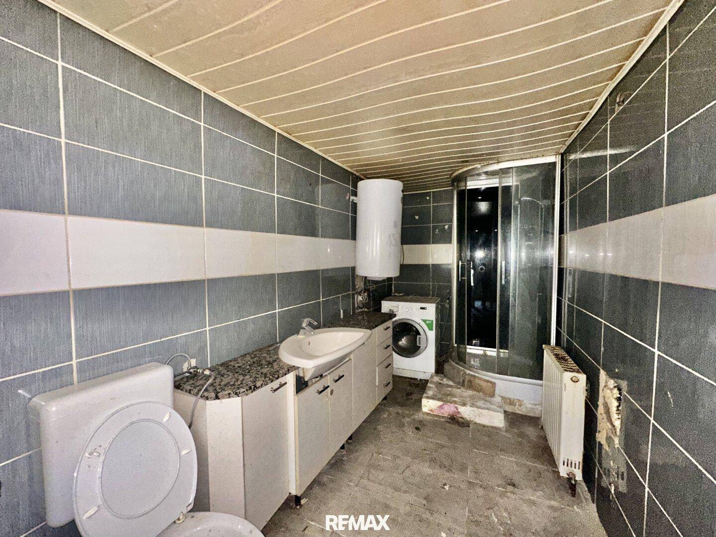 Badezimmer mit Dusche, Waschbecken und Toilette in sanierungsbedürftigem Zustand und alten Fliesen.