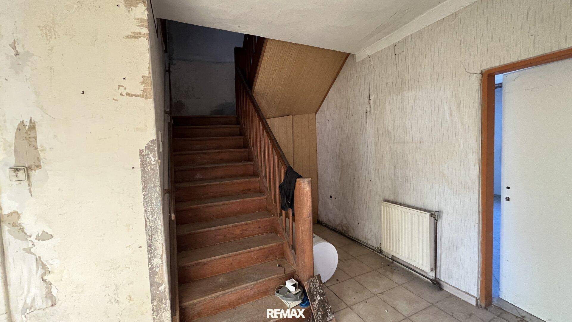 Alter Flur mit Holztreppe und abblätternder Tapete, der Renovierungsbedarf zeigt.