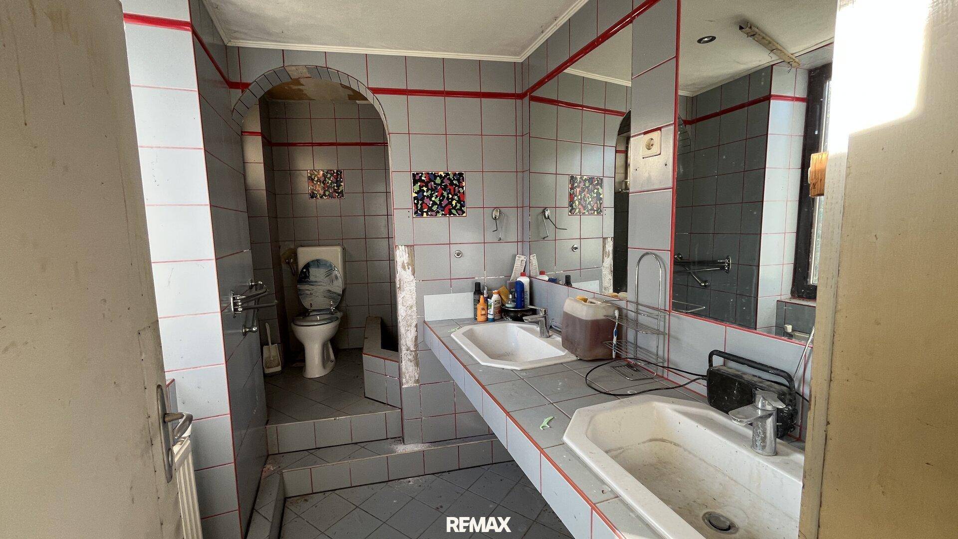 Veraltetes Badezimmer mit grauen Fliesen, roten Akzenten und zwei Waschbecken.