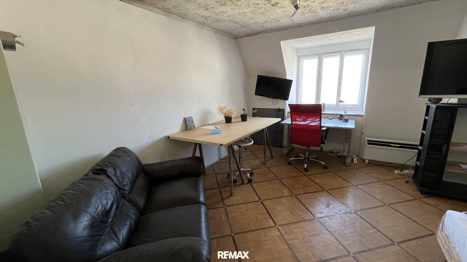 Wohnzimmer im Dachgeschoss mit schrägen Wänden, Fliesenboden und einem Arbeitsbereich.
