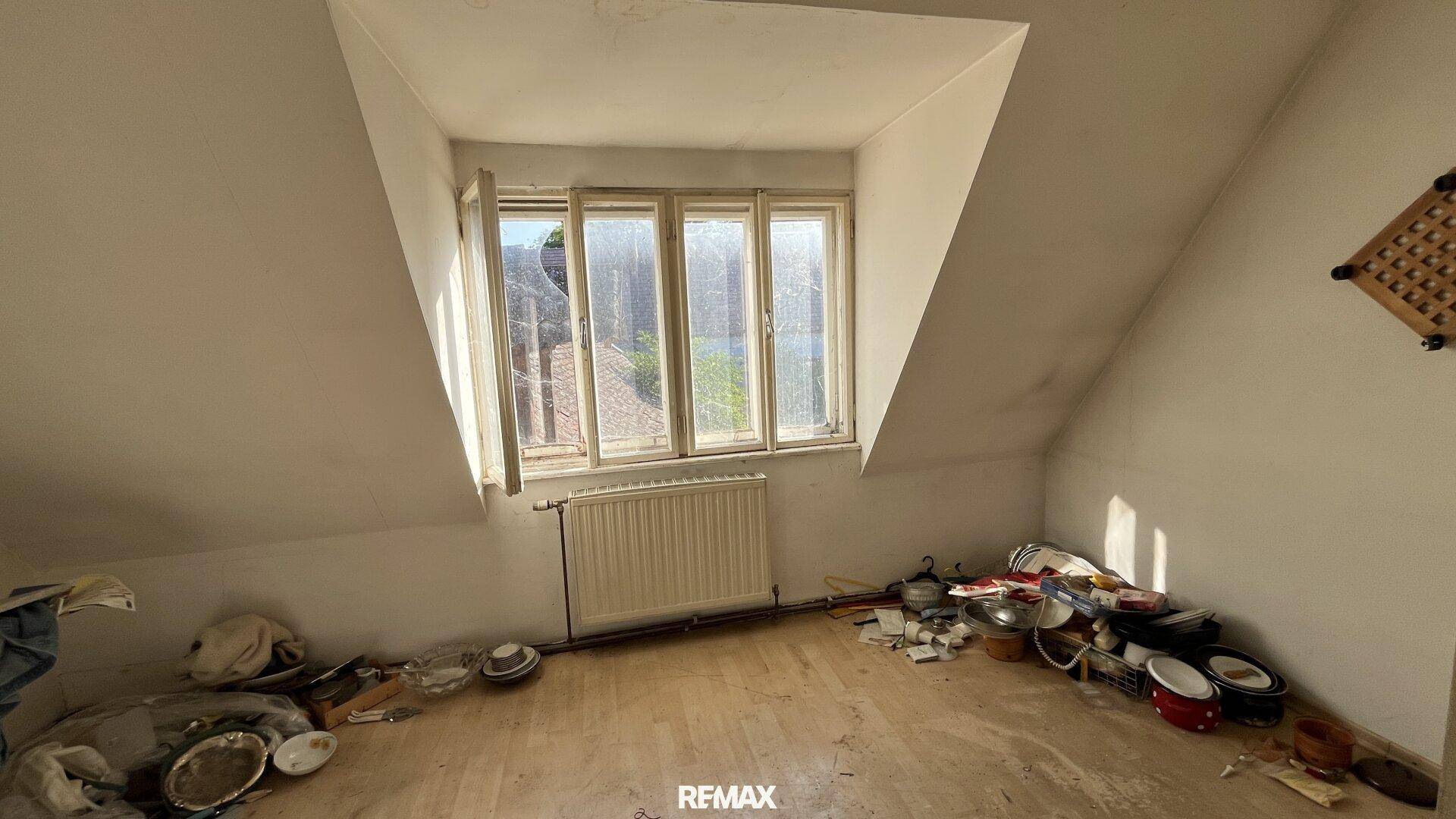 Leerer Raum im Dachgeschoss mit schrägen Wänden und einem Fenster, das Renovierung benötigt.