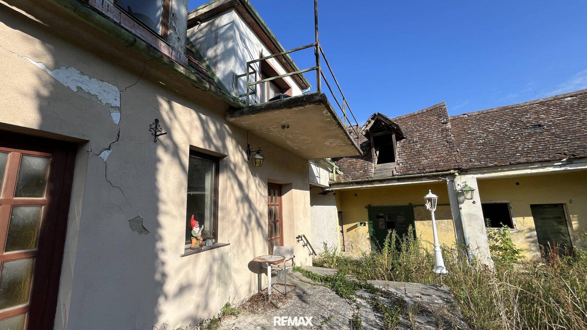 Altes, verfallenes Gebäude mit rissiger Fassade, überwuchertem Garten und kleinem Balkon.