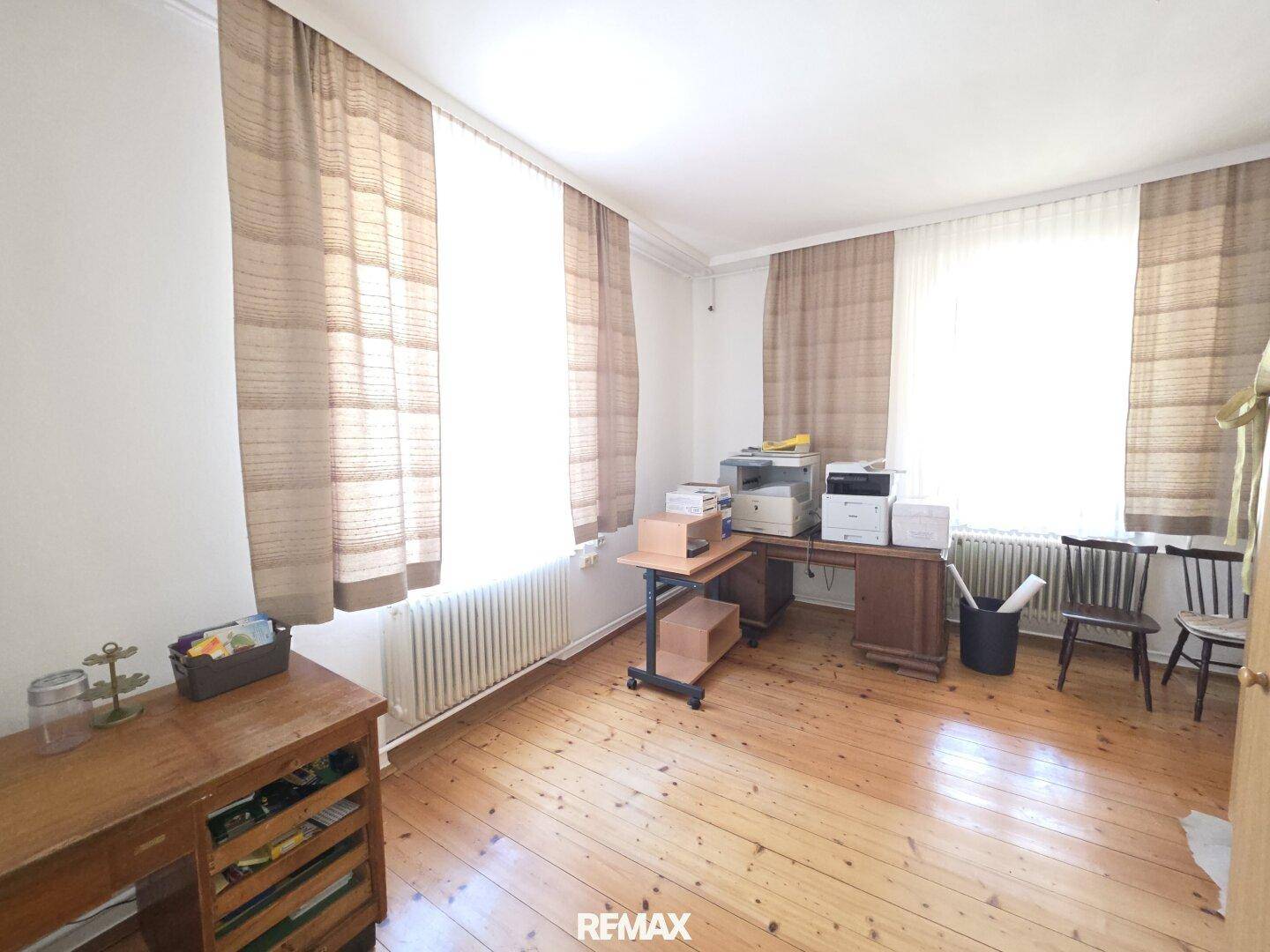 Helles Arbeitszimmer mit Holzdielenboden, zwei Fenstern und Büromöbeln.