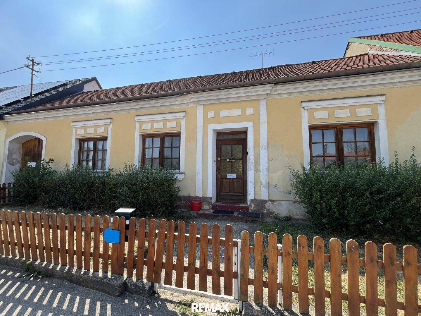 Vorderansicht eines traditionellen gelben Hauses mit Holzzaun und grünen Büschen.