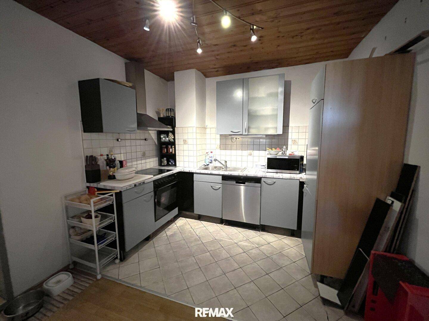 Funktionale Küche mit grauen Schränken, Fliesenboden und Holzdeckenverkleidung.