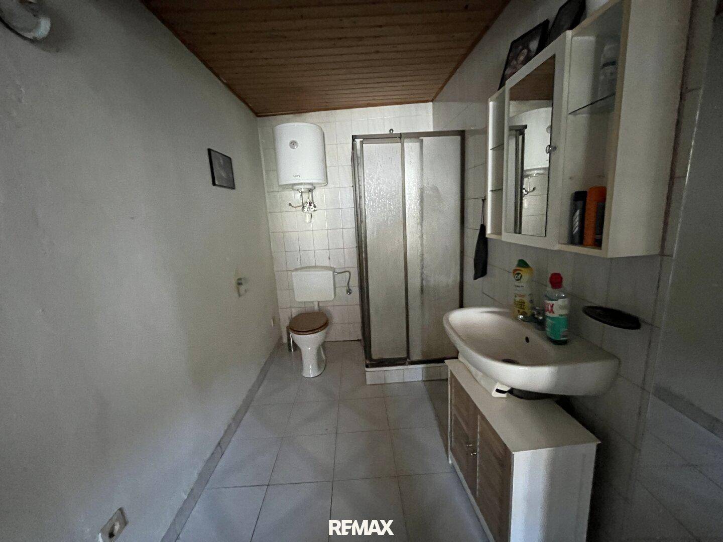 Helles Badezimmer mit Dusche, Waschbecken und weißen Fliesen, sowie Holzdeckenverkleidung.