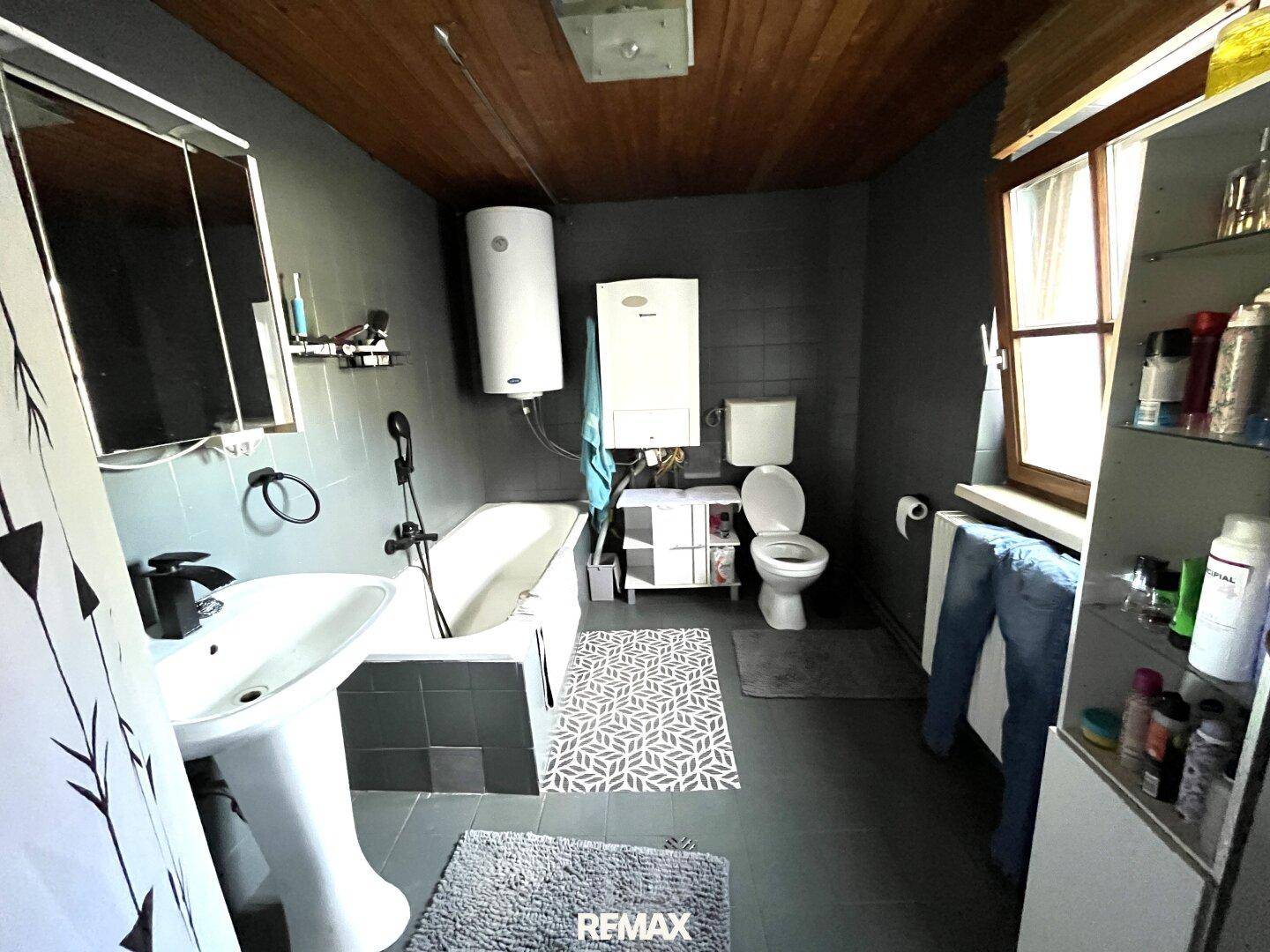 Modernes Badezimmer mit Badewanne, Waschbecken und dunklen Fliesen, sowie Holzdeckenverkleidung.