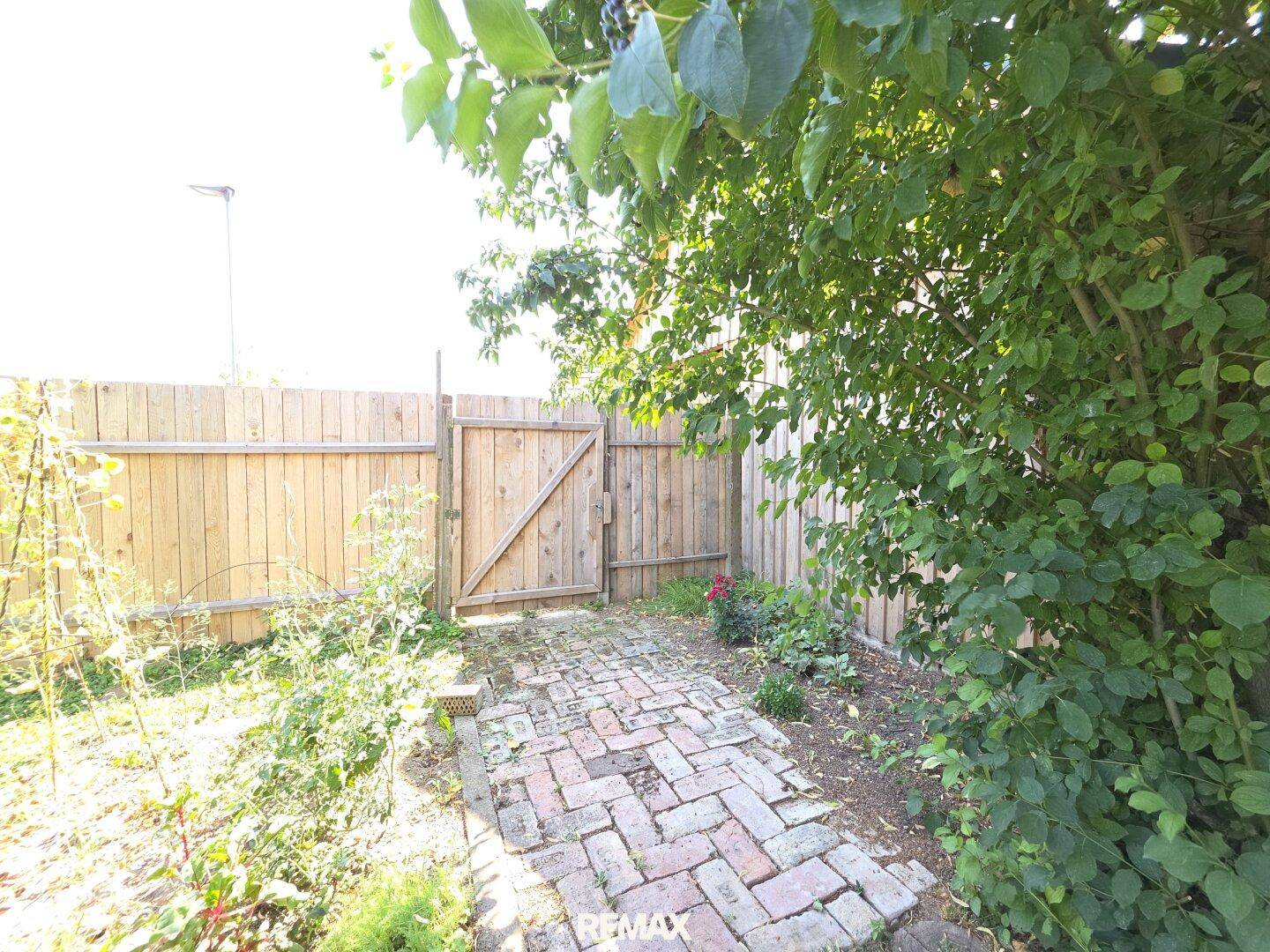 Rustikales Holztor in einem hohen Holzzaun, umgeben von Pflanzen und einem Ziegelsteinweg im Garten.