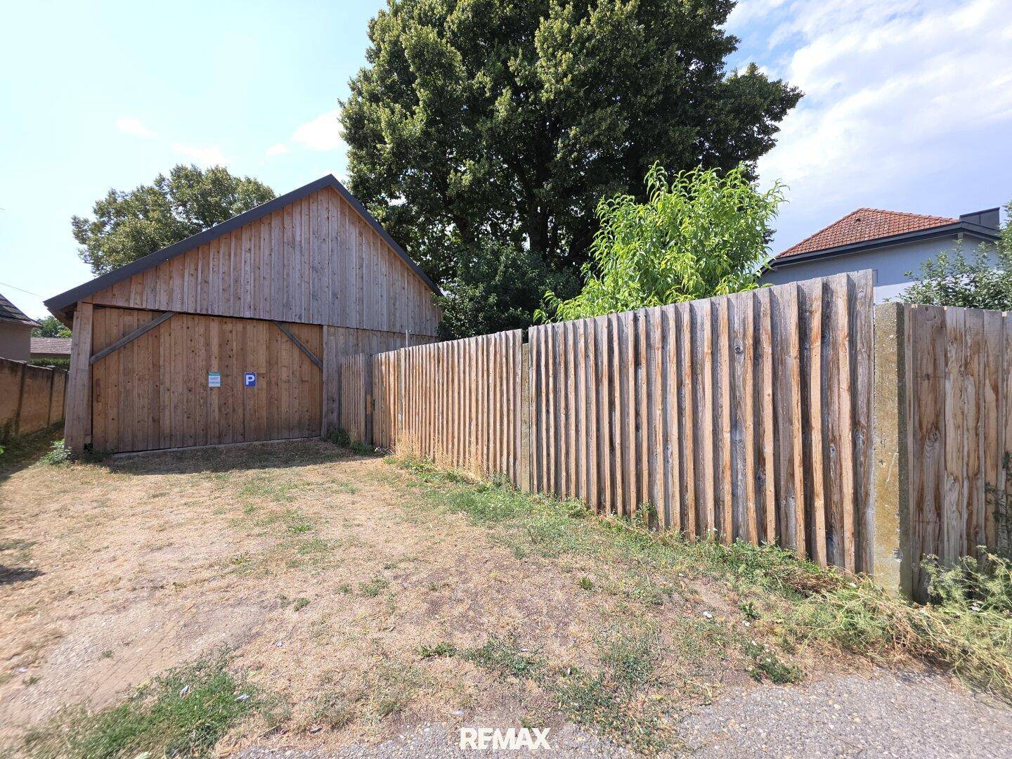 Vorderansicht eines Holzschuppens mit Parkplatz und einem robusten Holzzaun an der Grundstücksgrenze.