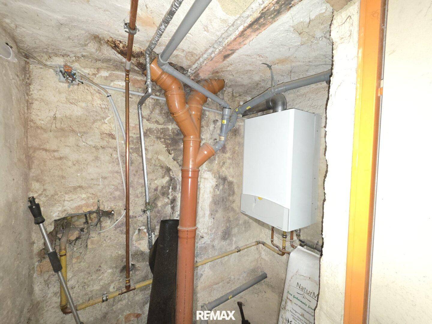 Kleiner Technikraum im Keller mit Wandheizkessel, sichtbaren Rohren und unverputzten Wänden.