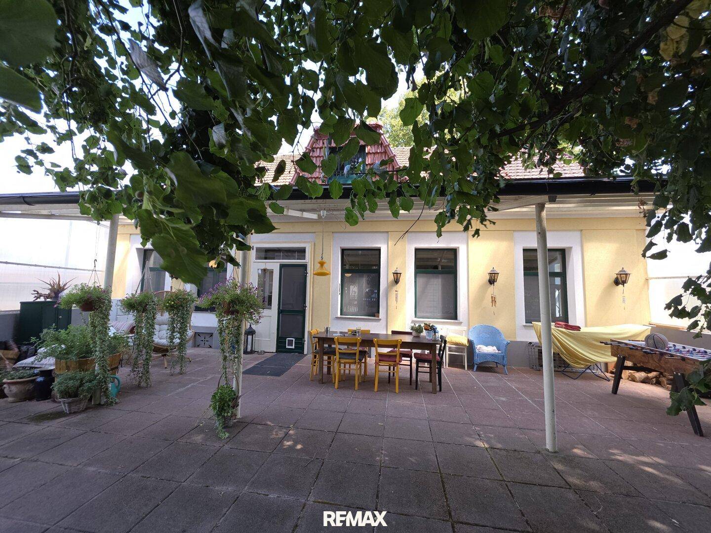 Große gepflasterte Terrasse mit Esstisch und Stühlen, umgeben von Bäumen und Pflanzen.