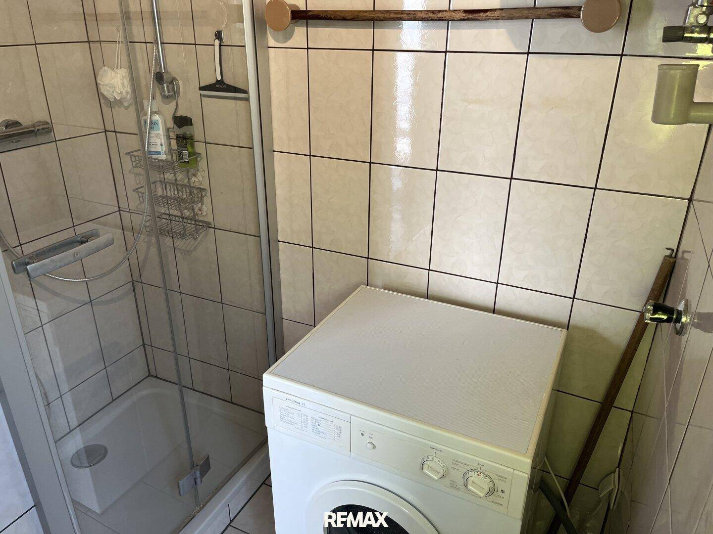 Badezimmer mit Duschkabine, Waschmaschine und hellen Fliesen an Wänden und Boden.