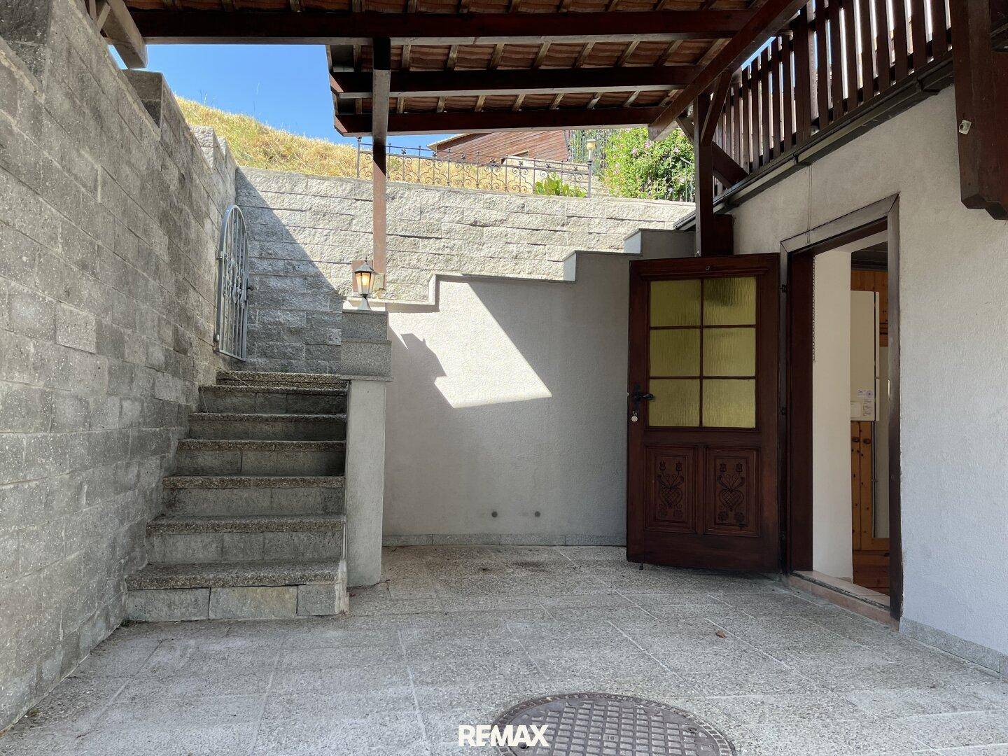 Gepflasterter Patio-Bereich unter einem Vordach mit Steintreppe und Zugang zum Haus.
