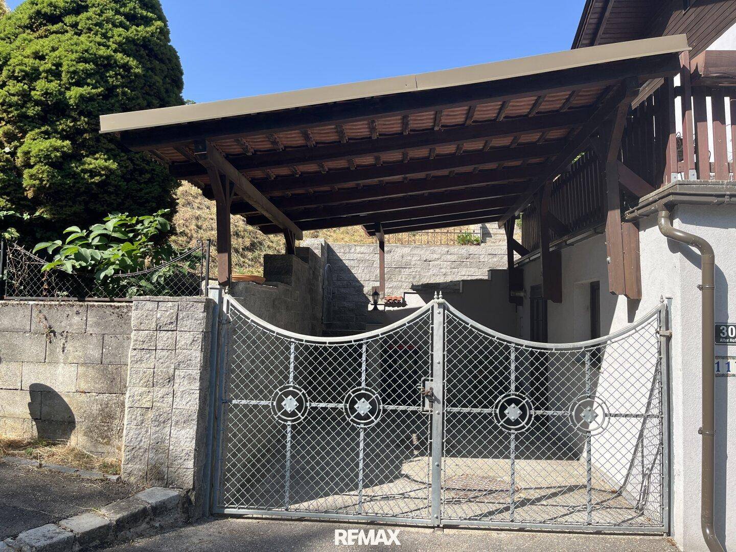 Einfahrt mit Metalltor und überdachtem Carport, umgeben von einer Steinmauer.