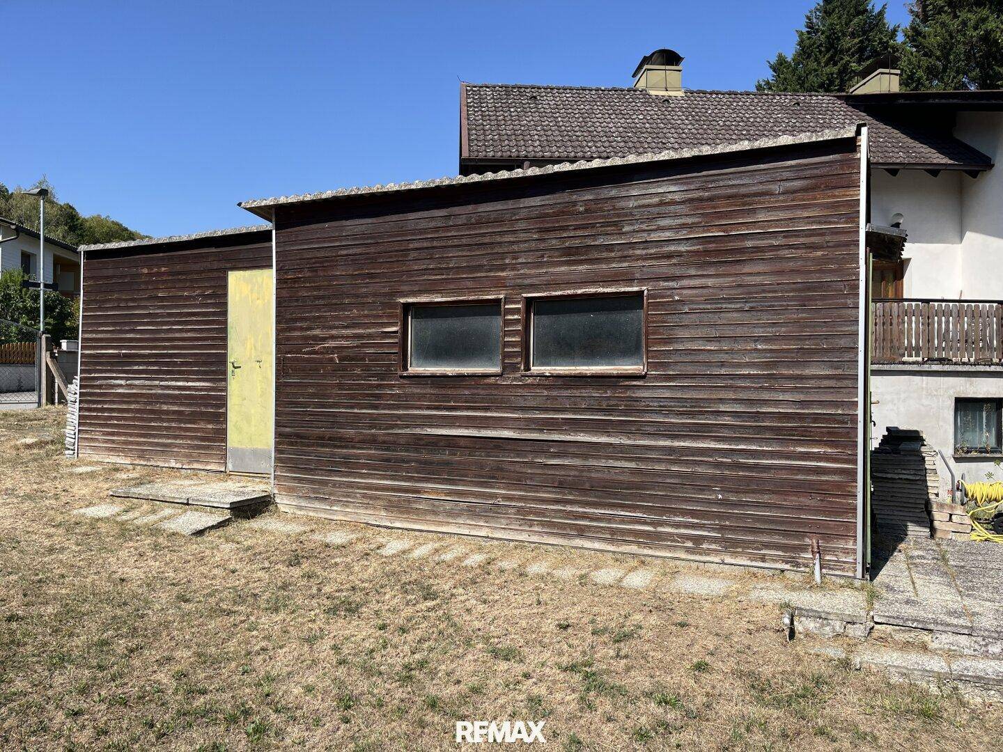 Rustikaler Holzschuppen mit zwei kleinen Fenstern auf einem trockenen Rasenbereich.