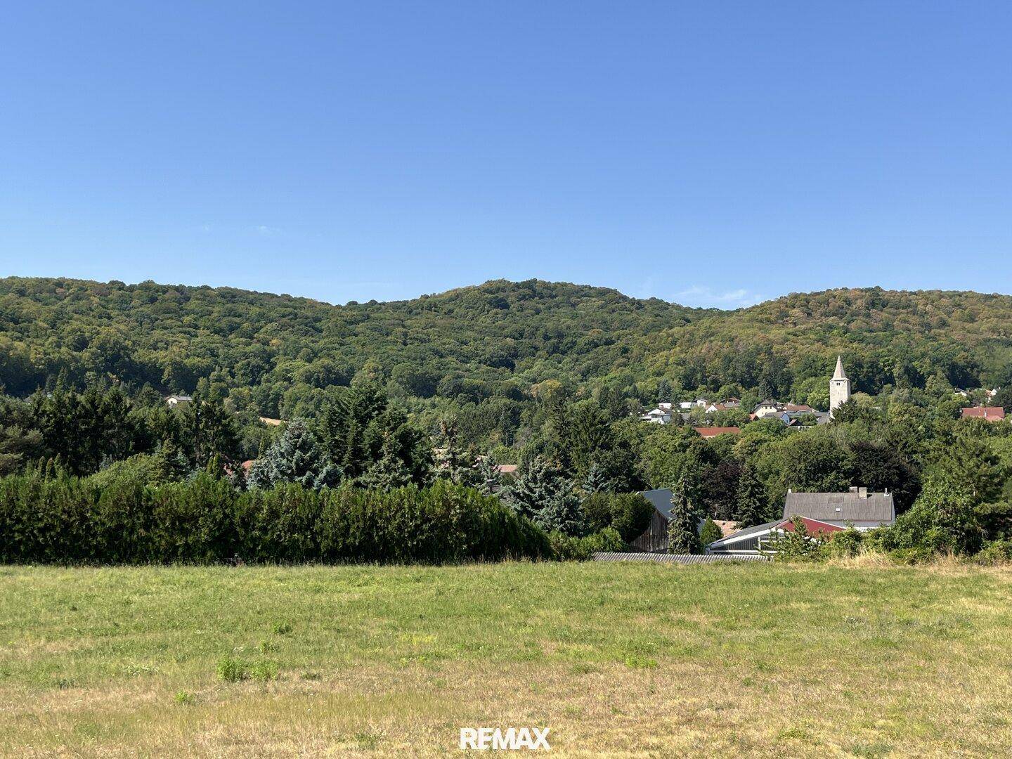 Weitläufiger Ausblick auf grüne Felder, eine bewaldete Hügellandschaft und ein Dorf mit Kirchturm.