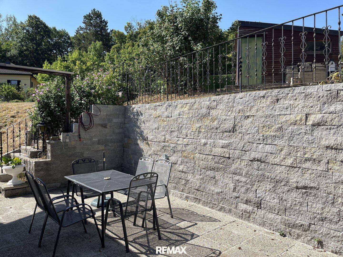 Sonniger Terrassenbereich mit Steinfliesen, Gartenmöbeln und einer Natursteinmauer.