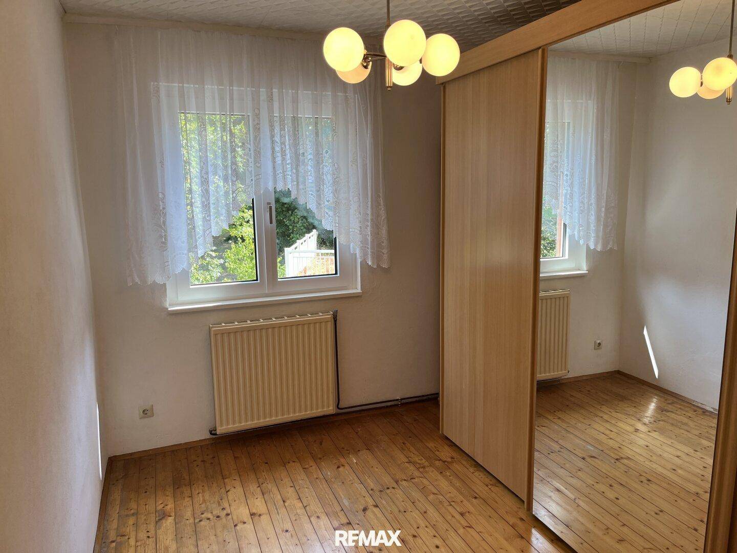 Helles Schlafzimmer mit Holzboden, Spiegelschrank und Fenster mit Blick ins Grüne.