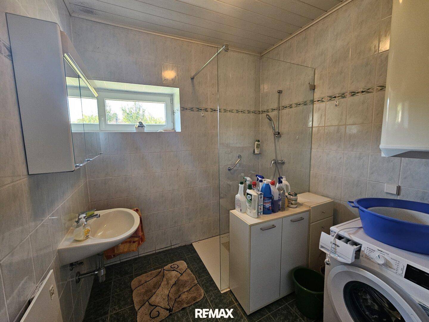 Badezimmer mit Dusche, Waschbecken, Waschmaschine und Fenster, das Tageslicht spendet.