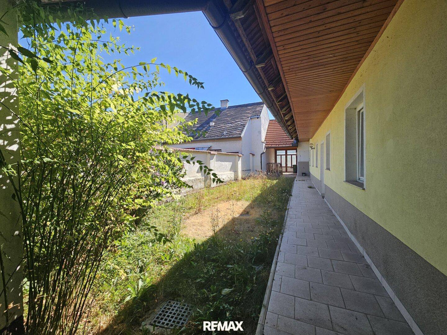 Schmaler Gartenstreifen entlang der Hauswand mit gepflastertem Weg und grüner Bepflanzung.
