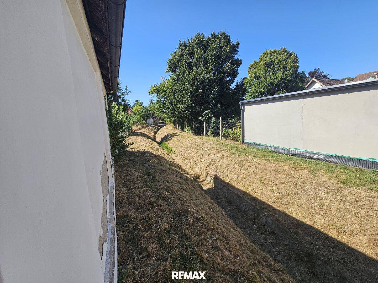 Entwässerungsgraben neben dem Grundstück mit trockener Vegetation und Bäumen im Hintergrund.