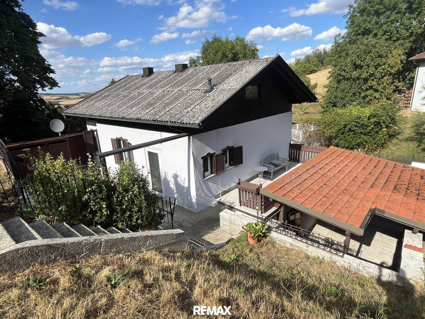Hausfassade mit Satteldach und kleiner Terrasse, umgeben von grüner Landschaft mit Fernblick.