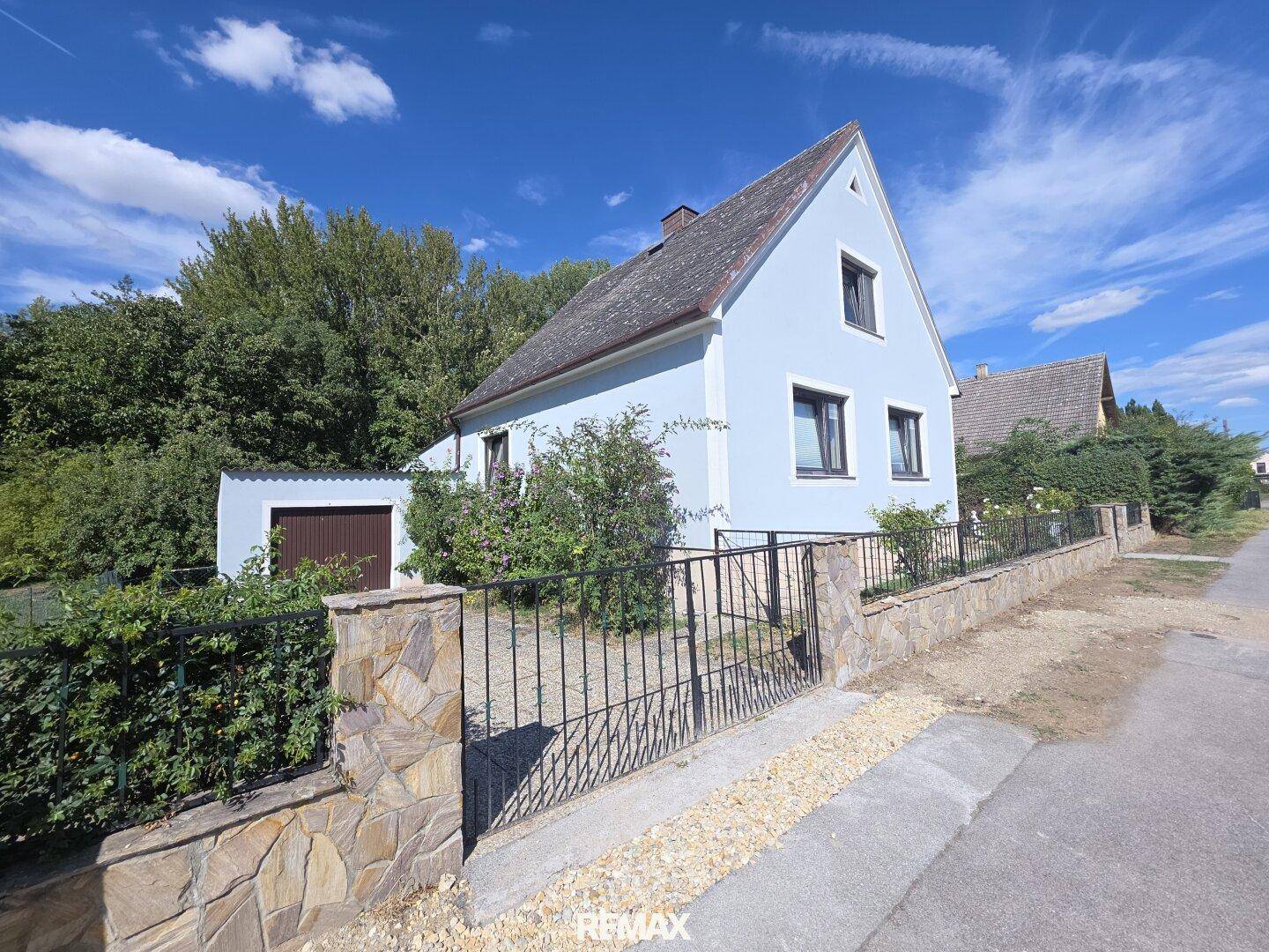 Einfamilienhaus mit hellblauer Fassade, Satteldach und Steinmauer, umgeben von grüner Vegetation.