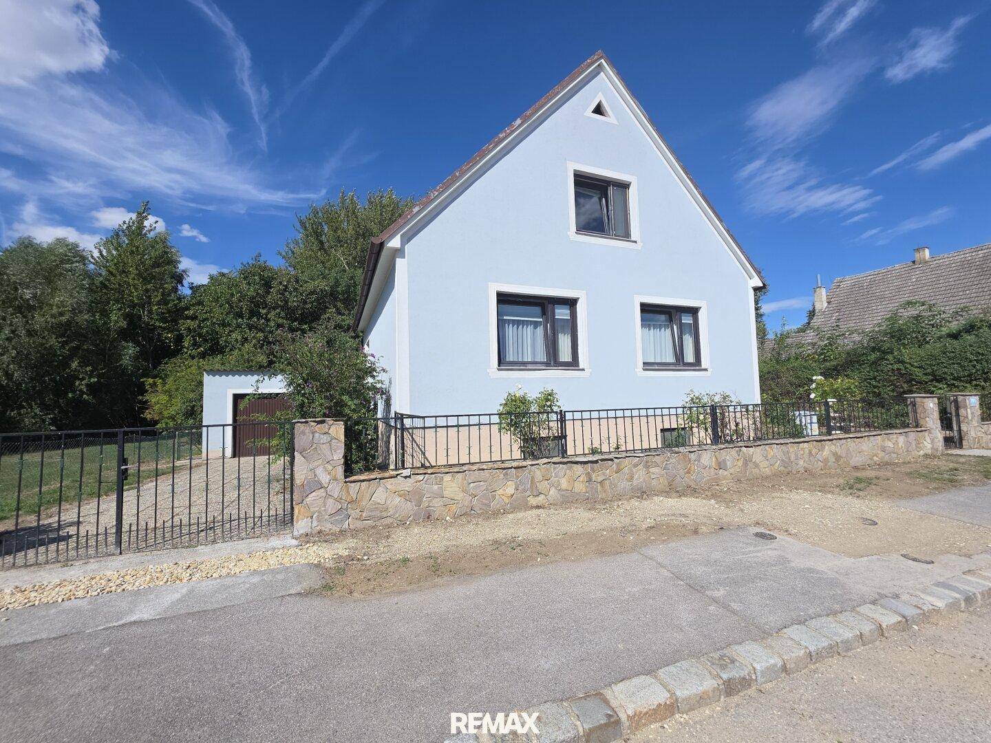 Freistehendes Haus mit hellblauer Fassade, Satteldach und gepflegtem Vorgarten unter blauem Himmel.