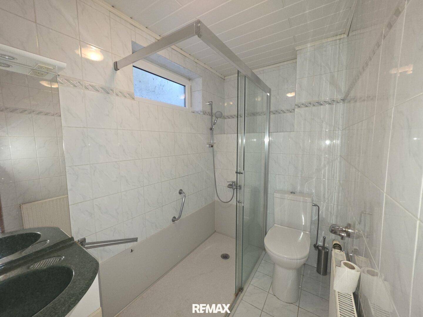 Badezimmer mit ebenerdiger Dusche, WC, Doppelwaschbecken und Haltegriffen für Barrierefreiheit.