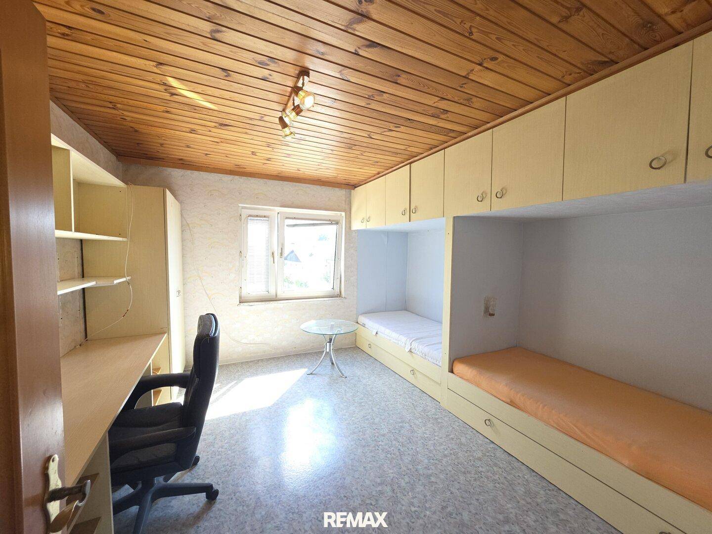 Kinderzimmer mit Etagenbetten, Schreibtisch und Holzdecke, hell durch Fenster.