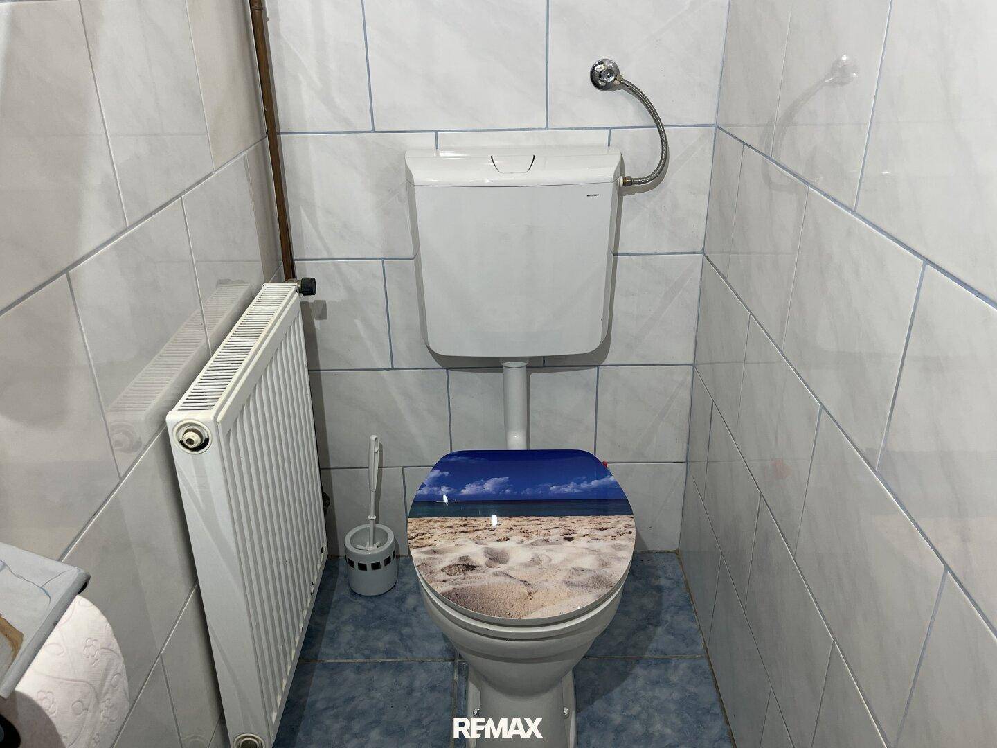 Separate Toilette mit Fliesenwänden, Heizkörper und einem dekorativen Toilettendeckel.