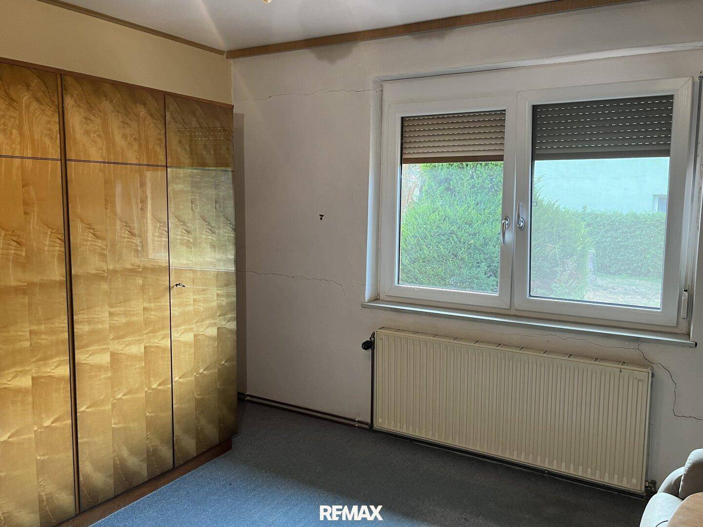 Schlafzimmer mit großem Holzschrank, Fenster mit Rollläden und sichtbaren Wandrissen.