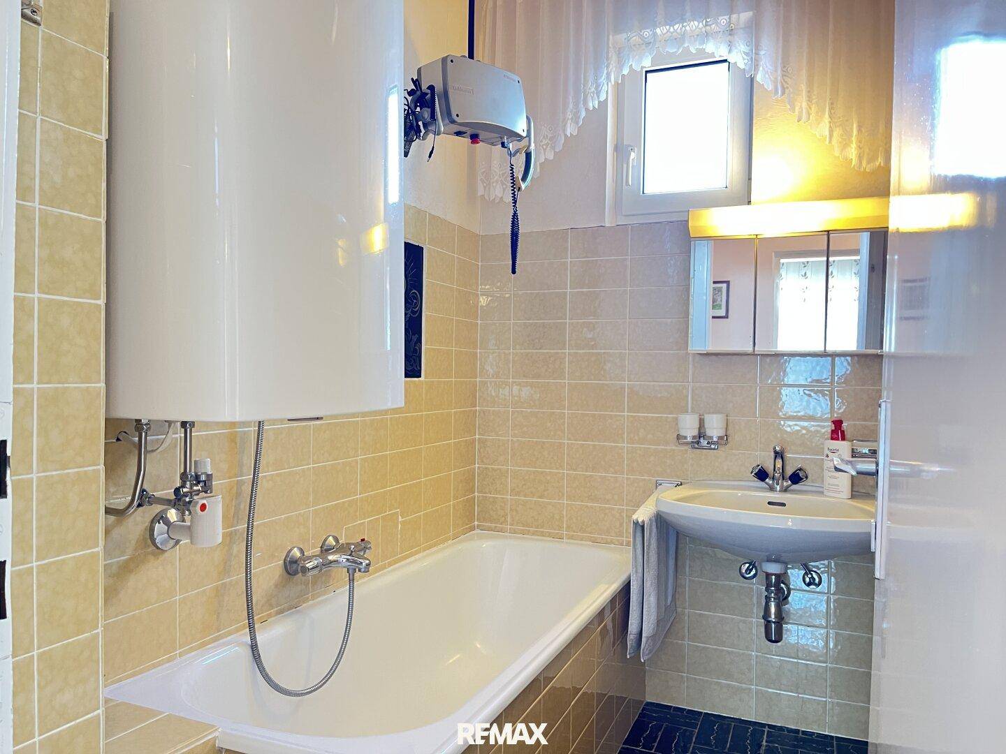 Badezimmer mit Badewanne, Waschbecken, Boiler und hellbraunen Wandfliesen.