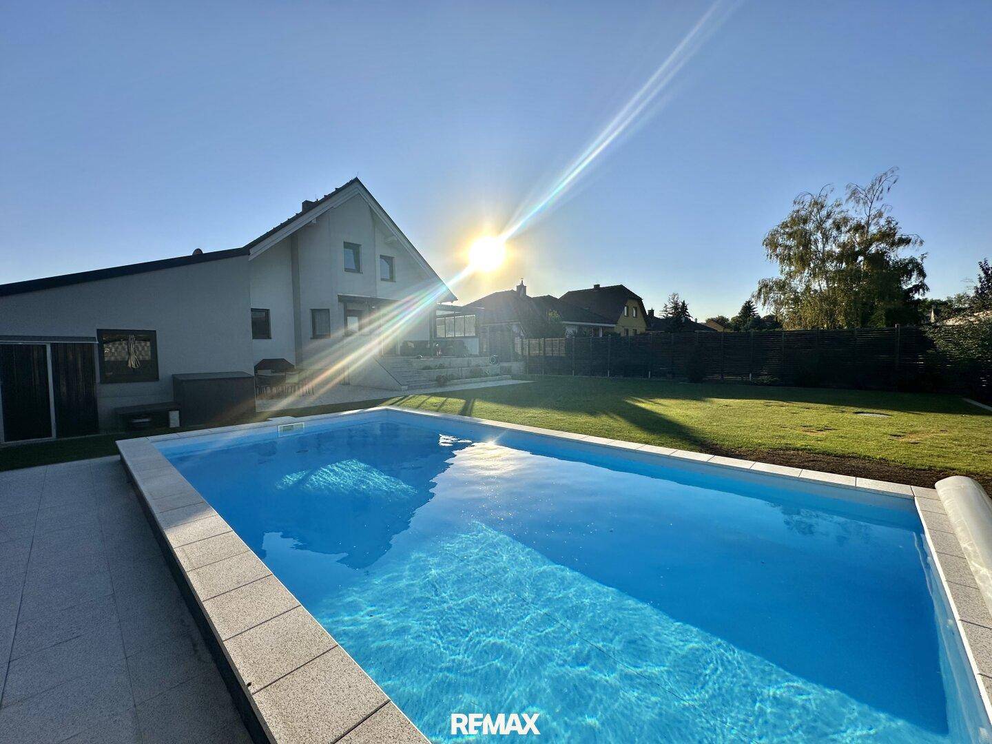 Sonniger Blick auf den Swimmingpool mit dem modernen Haus im Hintergrund und strahlendem Himmel.