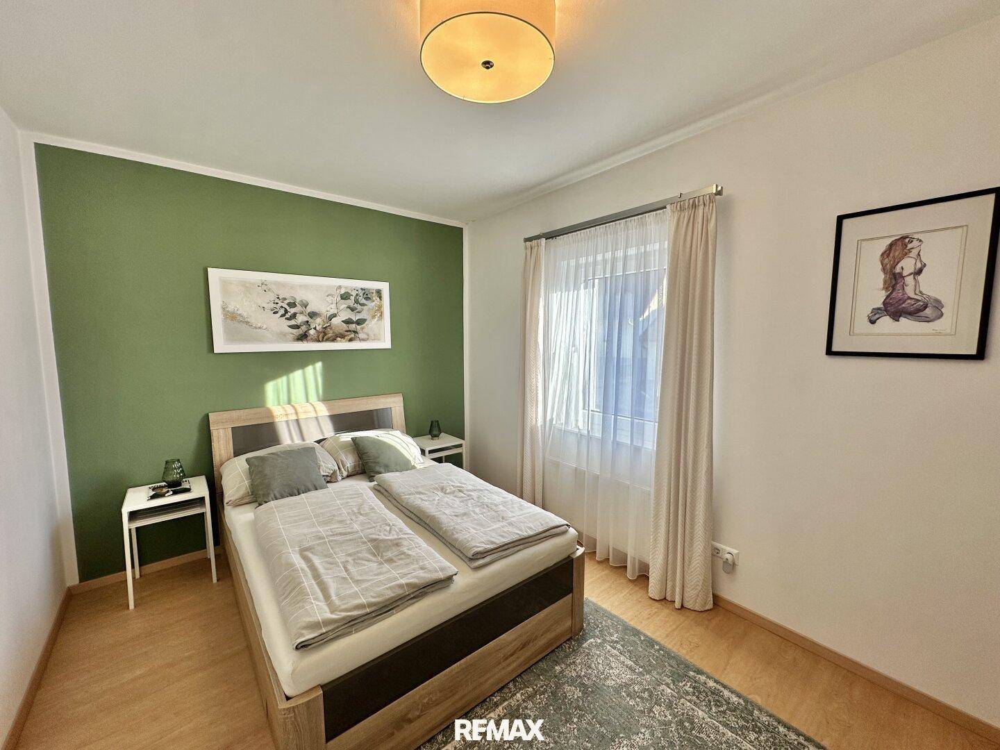 Gemütliches Schlafzimmer mit Doppelbett, grüner Akzentwand und Fenster mit Vorhängen.