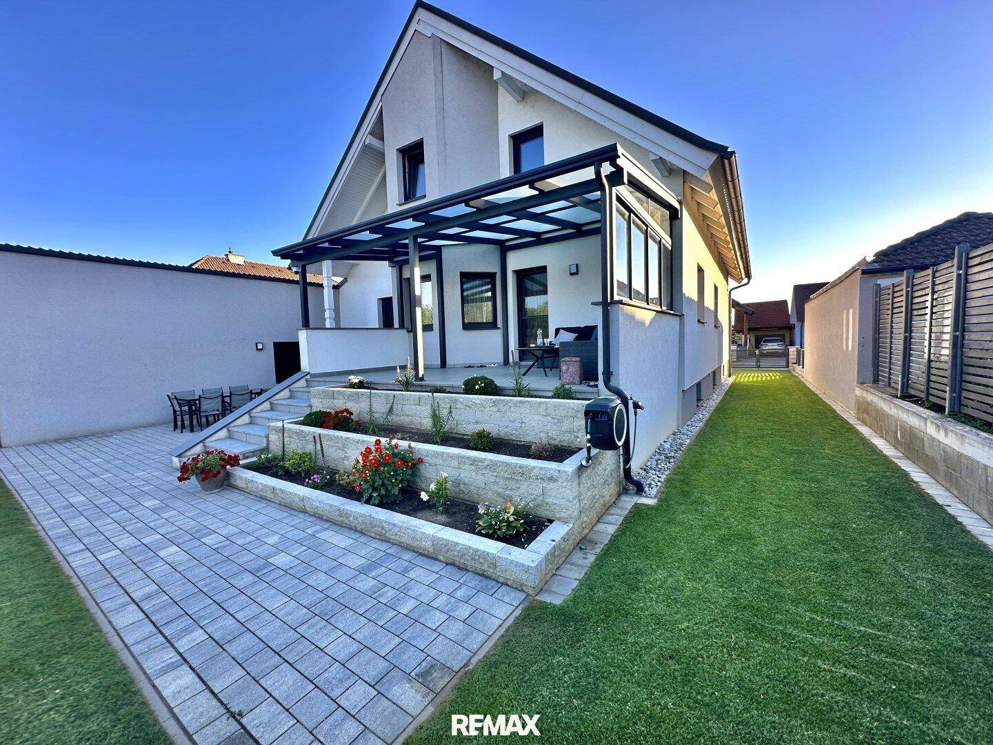 Modernes Einfamilienhaus mit überdachter Terrasse, gepflegtem Garten und gepflastertem Zugangsbereich.