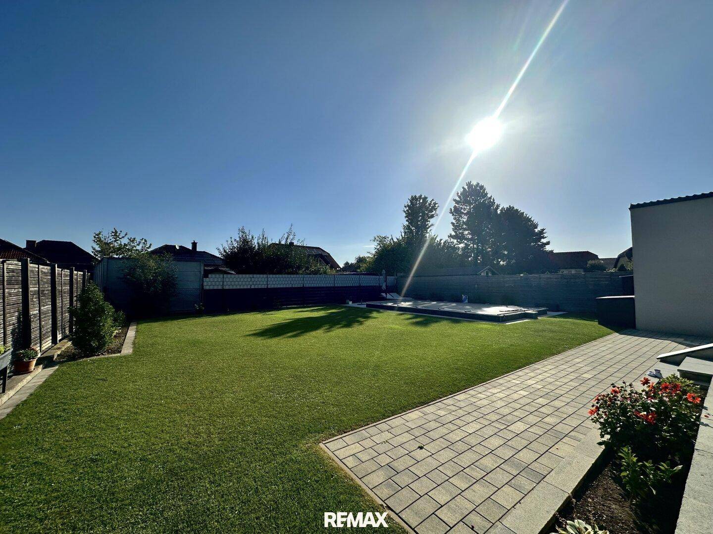 Sonniger Gartenbereich mit weitläufigem Rasen und gepflastertem Weg unter blauem Himmel.