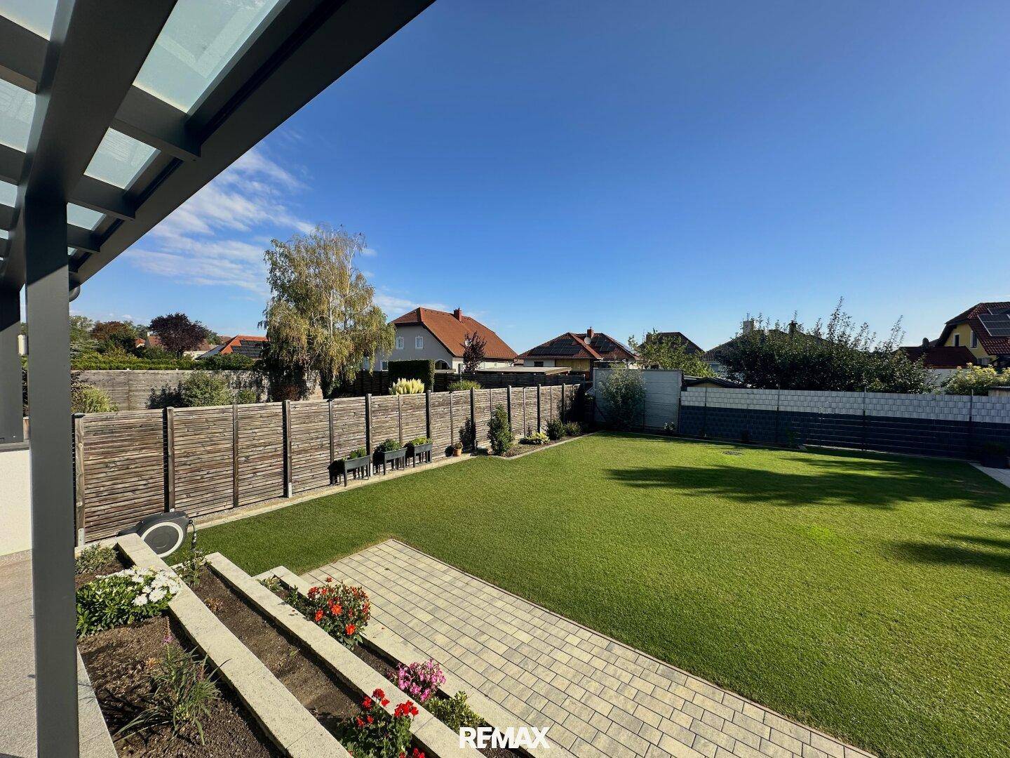 Blick von der überdachten Terrasse auf die Blumenbeete und den weitläufigen grünen Garten.