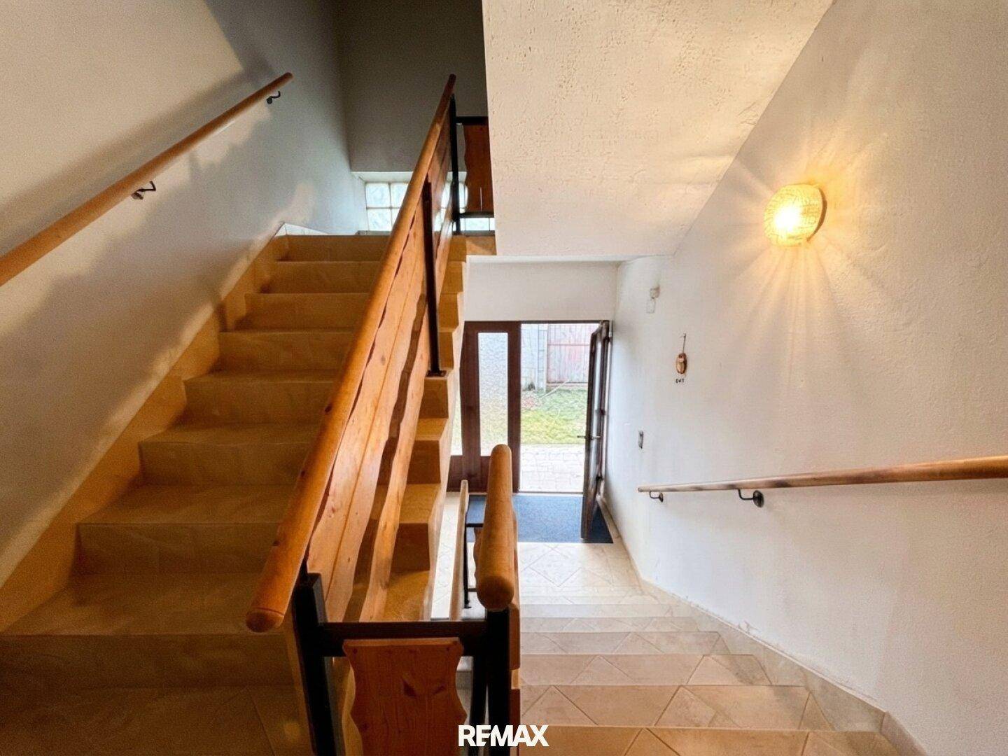 Treppenhaus mit Holzgeländer und gefliestem Boden, das zum Eingangsbereich führt.