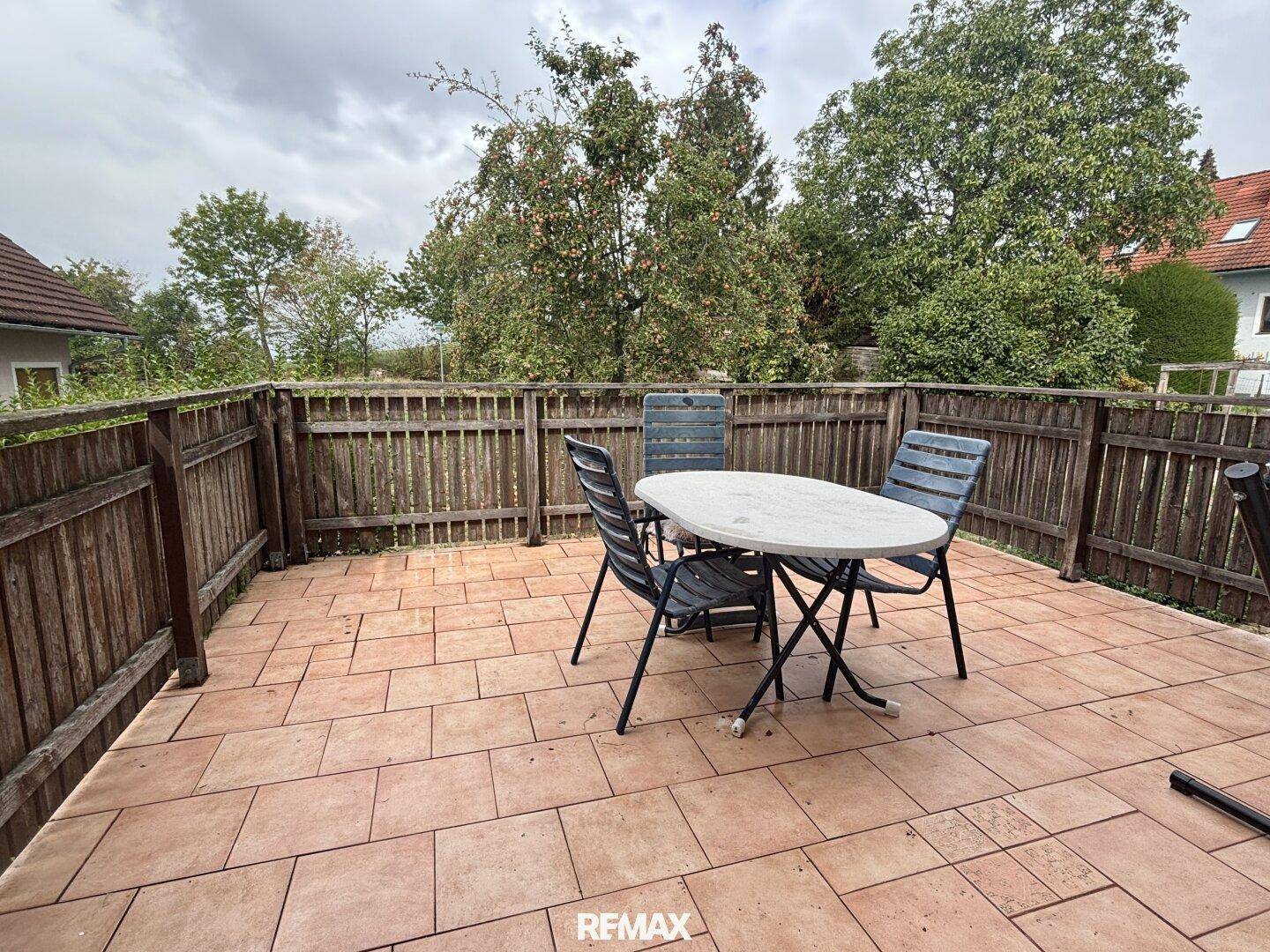 Große geflieste Terrasse mit Gartenmöbeln und Holzzaun, umgeben von Bäumen.