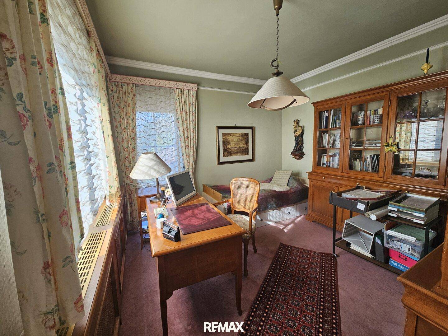 Arbeitszimmer mit Schreibtisch, Bücherregal und floralen Vorhängen, gut beleuchtet durch große Fenster.