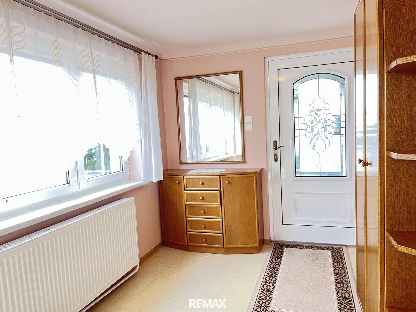 Heller Eingangsbereich mit Holzkommode, Spiegel und Fenster, das viel Tageslicht hereinlässt.