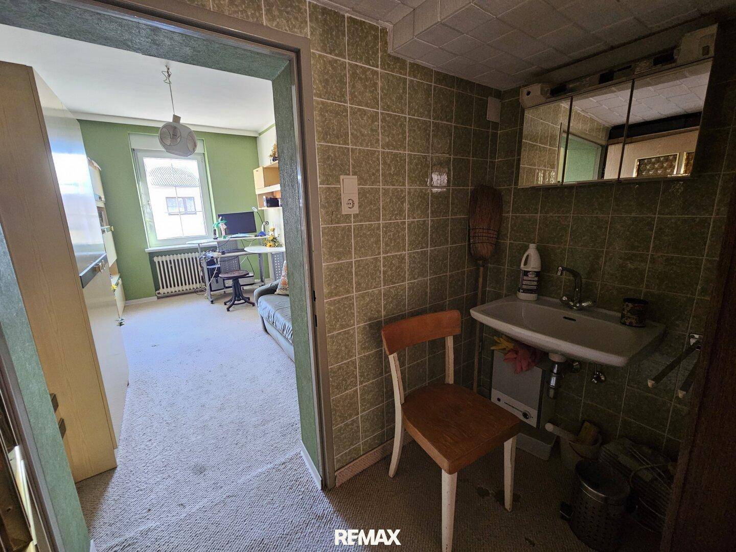 Kleiner Raum mit grün gefliesten Wänden, Waschbecken und Blick in ein angrenzendes Zimmer.