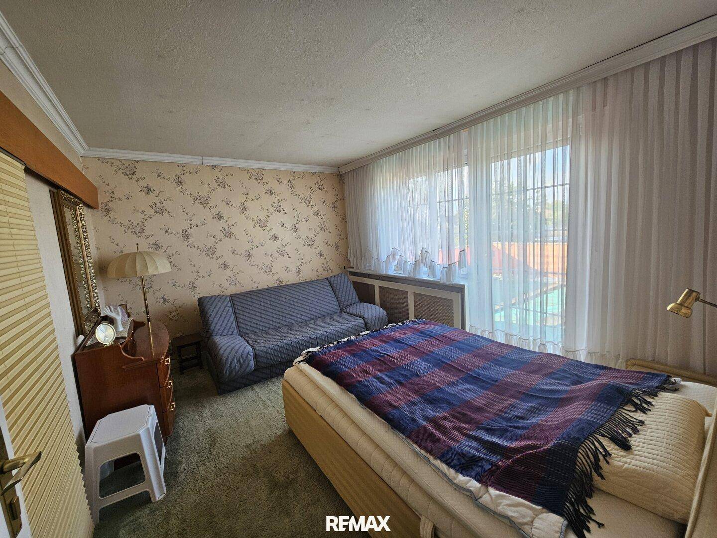 Schlafzimmer mit floraler Tapete, Teppichboden und großen Fenstern, die viel Licht hereinlassen.